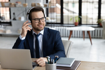 Distracted from computer work happy young successful businessman wearing modern headphones, enjoying pleasant music at workplace, relaxing in office, copy space for advertising no stress workday.