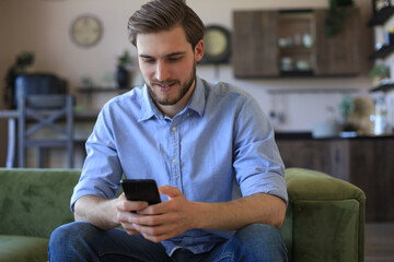 Attractive young man relaxing on a couch at home and using mobile phone for cheking social nets.