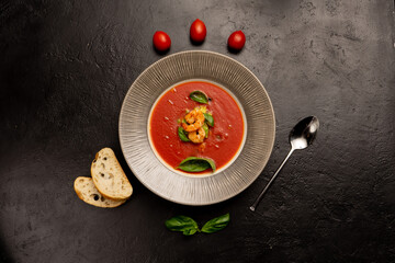 Classic Spanish tomato soup with prawns, basil leaves, cherry tomatoes, and ciabatta chunks on a black stone kitchen table.