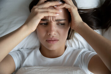 I did not get any sleep. Top view of exhausted unhappy millennial woman hugging head with bitter look lying in bed after sleepless night suffering of sudden migraine headache attack catching cold flu