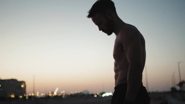 Silhouette of muscular bodybuilder with perfect body posing at the sunrise or sunset in a beach gym. Handsome strong man.