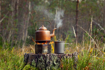 Vintage kettle on a tourist stove with a titanium mug on an old stump in the forest.