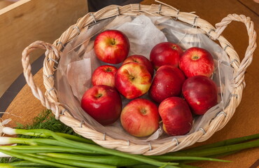 Red apples in a wicker basket and green onions close up on the background of the table in the kitchen