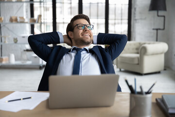 Happy young businessman in formal wear and eyeglasses stretching back with folded hands behind head, relaxing at modern workplace, finishing workday or meeting deadline, feeling no stress at work.