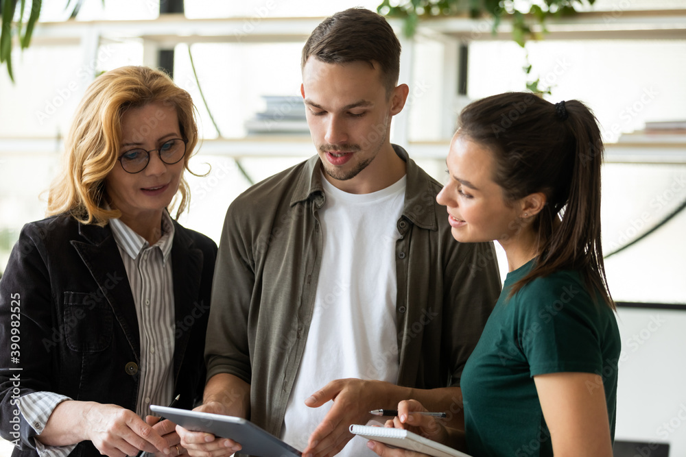 Canvas Prints Informal leader. Young man employee sharing information with women teammates using digital pad, diverse group of focused colleagues standing in office together discussing marketing strategy details