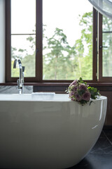 A delicate wedding bouquet of roses lies on the edge of a snow-white bathroom. Designer bathroom in the center of the bathroom. White artificial stone bathtub.