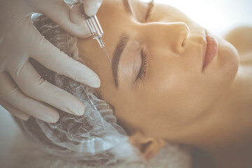 Beautician doing beauty procedure with syringe to face of young brunette woman in sunny clinic. Cosmetic medicine and surgery, beauty injections