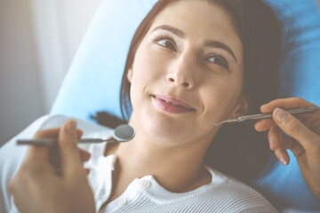 Smiling brunette woman being examined by dentist at dental clinic. Hands of a doctor holding dental instruments near patient's mouth. Healthy teeth and medicine concept