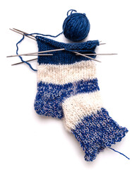 A half-knitted woolen sock in blue and white wool. Nearby are balls of woolen threads. Isolated on white background. Close-up. Knitting needles.