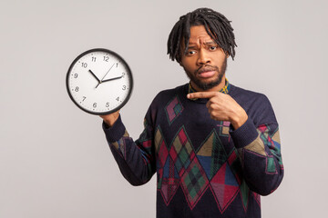 Nervous african man with beard and dreadlocks pointing finger on clock in his hand with serious...