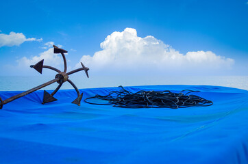 Rusty Anchor & black rope on a fishing boat covered in blue tarpaulin sheet on a beach with blue cloudy sky in the background