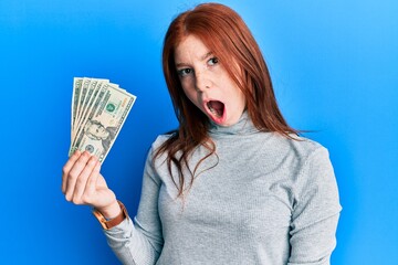 Young red head girl holding 20 dollars banknotes scared and amazed with open mouth for surprise, disbelief face