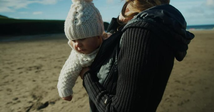 A young mother with her baby in a sling is standing on the beach in the autumn