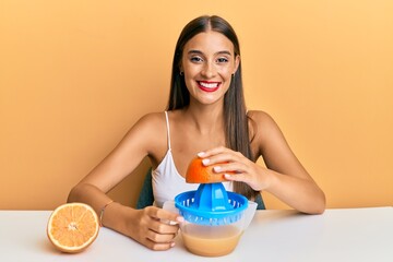 Young hispanic woman sitting on the table using juicer looking positive and happy standing and...
