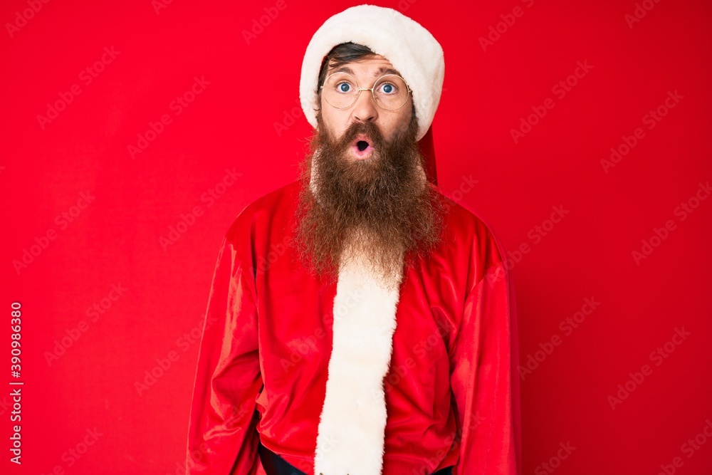 Wall mural Handsome young red head man with long beard wearing santa claus costume scared and amazed with open mouth for surprise, disbelief face