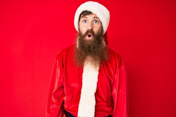 Handsome young red head man with long beard wearing santa claus costume scared and amazed with open mouth for surprise, disbelief face