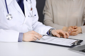 Fototapeta premium Unknown male doctor and patient woman discussing something while sitting in clinic and using clipboard. Best medical service in hospital, medicine, pandemic stop