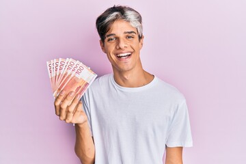 Young hispanic man holding 100 norwegian krone banknotes looking positive and happy standing and smiling with a confident smile showing teeth