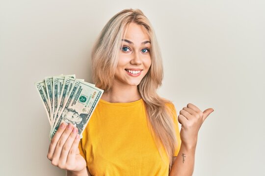 Beautiful caucasian blonde girl holding 20 dollars banknotes pointing thumb up to the side smiling happy with open mouth