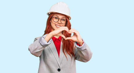Young redhead woman wearing architect hardhat smiling in love doing heart symbol shape with hands. romantic concept.