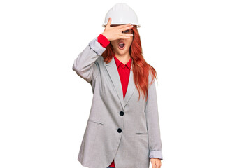 Young redhead woman wearing architect hardhat peeking in shock covering face and eyes with hand, looking through fingers with embarrassed expression.