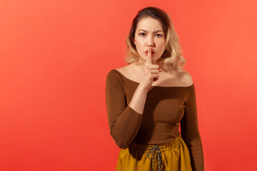 Shh, shut up! Serious and angry woman with blonde hair in brown casual blouse showing gesture secret sign with finger near her lips. Expression emotion. Indoor isolated on red background