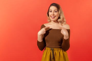 Portrait of charming happy woman in casual blouse and trousers smiling and pointing at herself, being proud of own success, boasting of achievement. Indoor studio shot isolated on red background