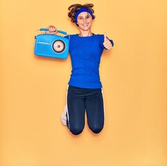 Young beautiful sportswoman listening to music smiling happy. Jumping with smile on face holding vintage radio doing ok sign with thumbs up over isolated yellow background.