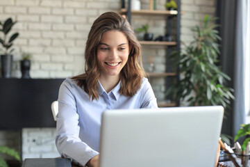 Smiling pretty woman sitting at table, looking at laptop screen. Happy entrepreneur reading message email with good news, chatting with clients online.