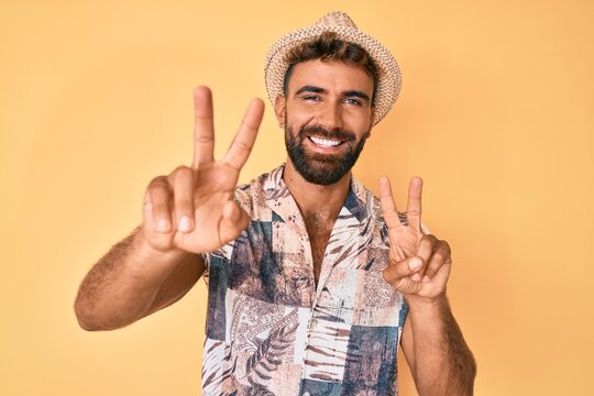 Young hispanic man wearing summer hat smiling looking to the camera showing fingers doing victory sign. number two.
