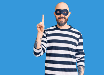 Young handsome man wearing burglar mask pointing finger up with successful idea. exited and happy. number one.