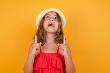 young Caucasian girl standing against yellow background being amazed and surprised looking and pointing up with fingers showing something strange.