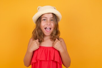 Young Caucasian little girl wearing hat and red dress against yellow rejoicing success and victory clenching his fists with joy being happy to achieve her aim and goals. Positive emotions, feelings.
