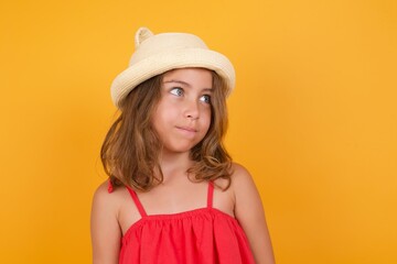 Young Caucasian little girl wearing hat and red dress against orange background, looks pensively aside, plans actions after school, imagines what to do Thinks over about new project. 
