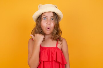 Shocked young Caucasian girl standing against yellow background  points with thumb away, indicates something. Check this out. Advertisement concept.
