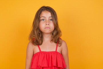 Dreamy young Caucasian girl standing against yellow background keeps hands pressed together under chin, looks with happy expression, has toothy smile.