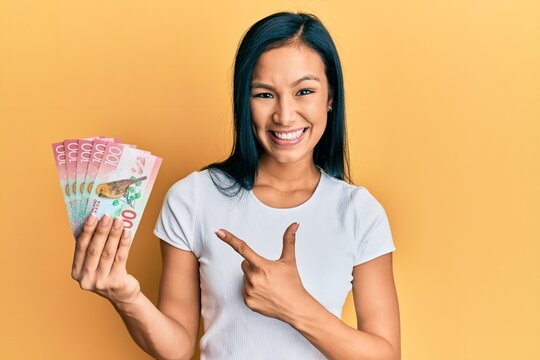 Beautiful hispanic woman holding 100 new zealand dollars banknote smiling happy pointing with hand and finger