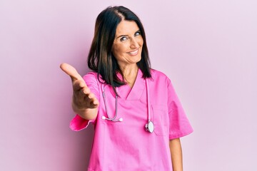 Middle age brunette woman wearing doctor uniform and stethoscope smiling friendly offering...