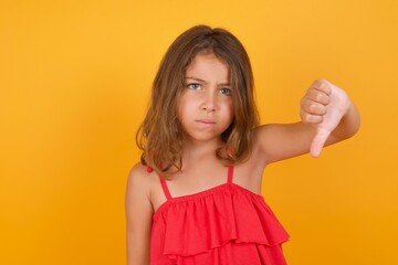 Discontent Caucasian young girl standing against yellow background shows disapproval sign, keeps thumb down, expresses dislike, frowns face in discontent. Negative feelings.