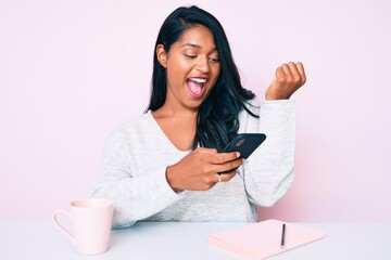Beautiful latin young woman with long hair using smartphone sitting on the table screaming proud, celebrating victory and success very excited with raised arms
