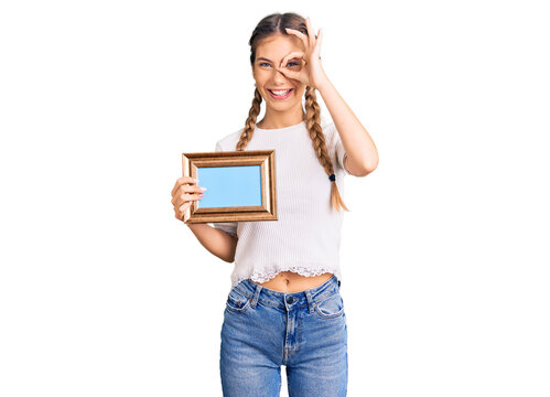 Beautiful caucasian woman with blonde hair holding empty frame smiling happy doing ok sign with hand on eye looking through fingers