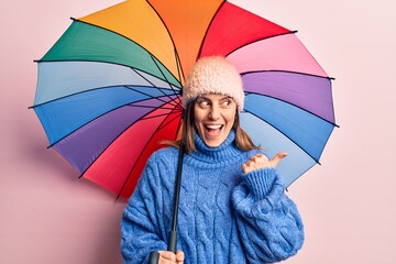 Young beautiful woman holding colorful umbrella pointing thumb up to the side smiling happy with open mouth