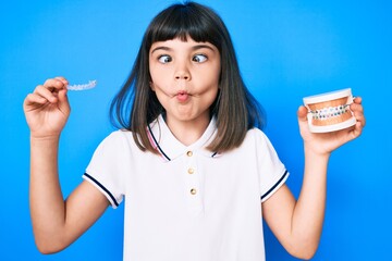 Young little girl with bang holding invisible aligner orthodontic and braces making fish face with...