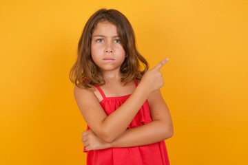 young Caucasian girl standing against yellow background smiling broadly at camera, pointing fingers away, showing something interesting and exciting.