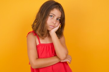Very bored young Caucasian girl standing against yellow background holding hand on cheek while support it with another crossed hand, looking tired and sick.