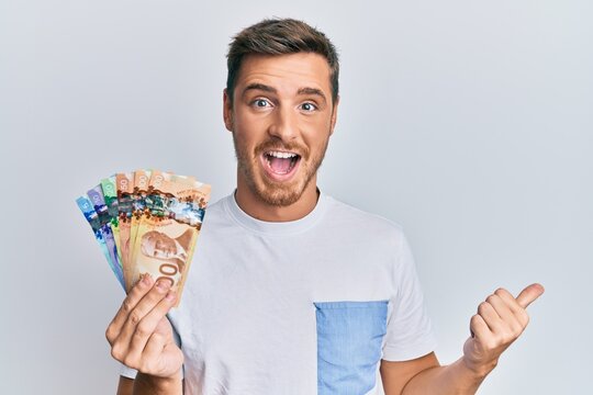 Handsome caucasian man holding canadian dollars pointing thumb up to the side smiling happy with open mouth
