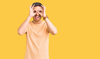 Handsome young man with bear wearing casual yellow tshirt doing ok gesture like binoculars sticking tongue out, eyes looking through fingers. crazy expression.