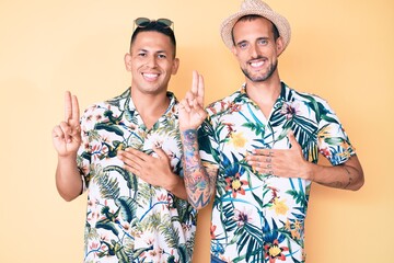 Young gay couple of two men wearing summer hat and hawaiian shirt smiling swearing with hand on...