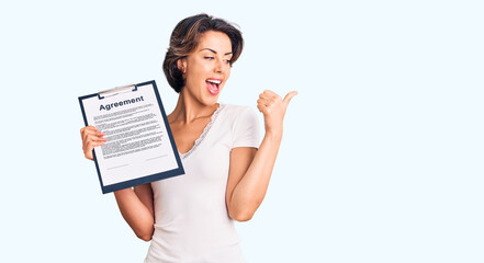 Young beautiful woman holding clipboard with agreement document pointing thumb up to the side smiling happy with open mouth