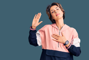 Young beautiful woman wearing sportswear swearing with hand on chest and open palm, making a...